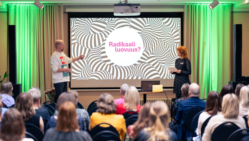 Kuvituskuva, jossa kaksi puhujaa esitelmöivät luovuudesta lavalla. Yleisö seuraa esitystä ja osallistuu keskusteluun.